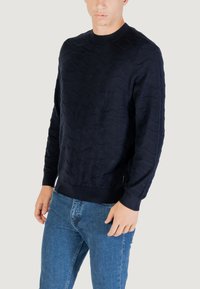 Dunkelblauer Pullover mit strukturiertem Wellenmuster, Rundhalsausschnitt, und gerippten Bündchen sowie Saum. Dazu werden hellblaue Denim-Jeans getragen. Mittelschwere Strickware.