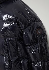 Chaqueta acolchada azul marino brillante con secciones acolchadas, un parche circular con logo en el hombro y textura brillante en toda la prenda.