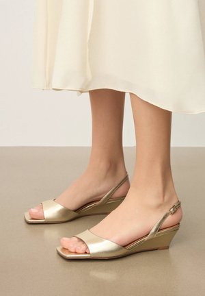 Picioare purtând sandale cu platformă cu bretea aurie metalizată și degete rotunde, asortate cu o fustă fluidă de culoare crem.