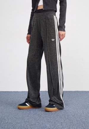 Osoba nosząca czarne błyszczące spodnie dresowe z białymi paskami i logo Adidas, w połączeniu z ciemnymi butami z gumowymi podeszwami i krótką czarną kurtką.