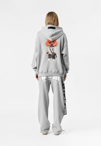 Persona vistiendo un conjunto de sudadera y pantalones grises, la sudadera presenta un gran gráfico de un oso de peluche en la parte trasera, de pie contra un fondo blanco liso.