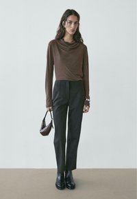 Brązowy, dłoni rękaw top w stylu drapowanym zestawiony z czarnymi, wysokim stanem spodniami. Modelka trzyma małą brązową torebkę. Czarnie botki na kostkę dopełniają stylizację.