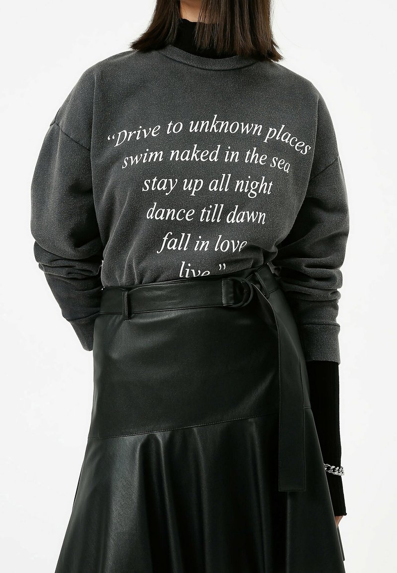 Sweat-shirt gris avec texte blanc et manches longues, assorti à une jupe en cuir noire avec une taille ceinturée et un design fluide.
