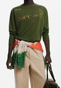 Maglietta a maniche lunghe verde oliva con testo stampato. Sciarpa a righe crema e arancione legata in vita. Pantaloni beige. Tiene una borsa verde.