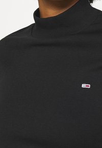 Svart polotröja i mjukt stickat tyg med en liten röd, vit och blå logga broderad på bröstet.