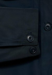Detalle de chaqueta azul marino con una textura de tela suave, dos botones negros y un dobladillo doblado con costuras precisas.