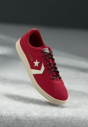 Red suede sneaker with black laces, white star logo, and beige sole. Features a low-top design and a smooth texture.