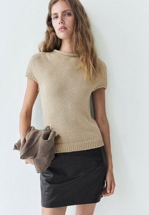 Frau mit langen Haaren, die ein beiges kurzärmeliges Stricktop und einen schwarzen Lederrock trägt und eine braune Wildlederjacke vor weißem Hintergrund hält.