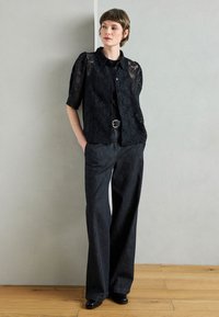 Blusa corta in pizzo nero con collo a camicia, abbinata a pantaloni larghi in denim scuro e scarpe nere. Sfondo in legno.
