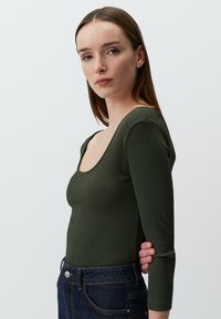Body a maniche lunghe a coste verde oliva con scollo rotondo, abbinato a jeans in denim scuro. Il tessuto è liscio e aderente.