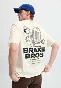 Cremfarvet t-shirt med et tegneseriegraphic af en mekaniker med værktøj, et dæk og fed sort tekst "BRAKE BROS" på ryggen.