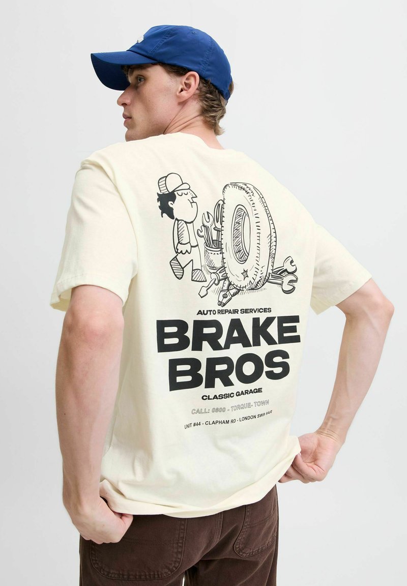 Cremfarvet t-shirt med et tegneseriegraphic af en mekaniker med værktøj, et dæk og fed sort tekst "BRAKE BROS" på ryggen.