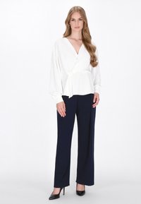 Blusa avvolgente bianca con maniche lunghe, abbinata a pantaloni larghi blu navy. L'outfit presenta un tessuto morbido e una silhouette favorevole. Tacchi neri.
