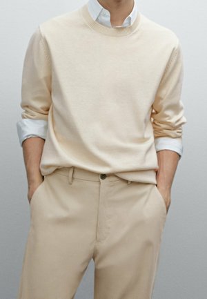 Personne portant un pull beige clair sur une chemise blanche avec les manches relevées et un pantalon beige assorti, mains dans les poches devant un fond uni.