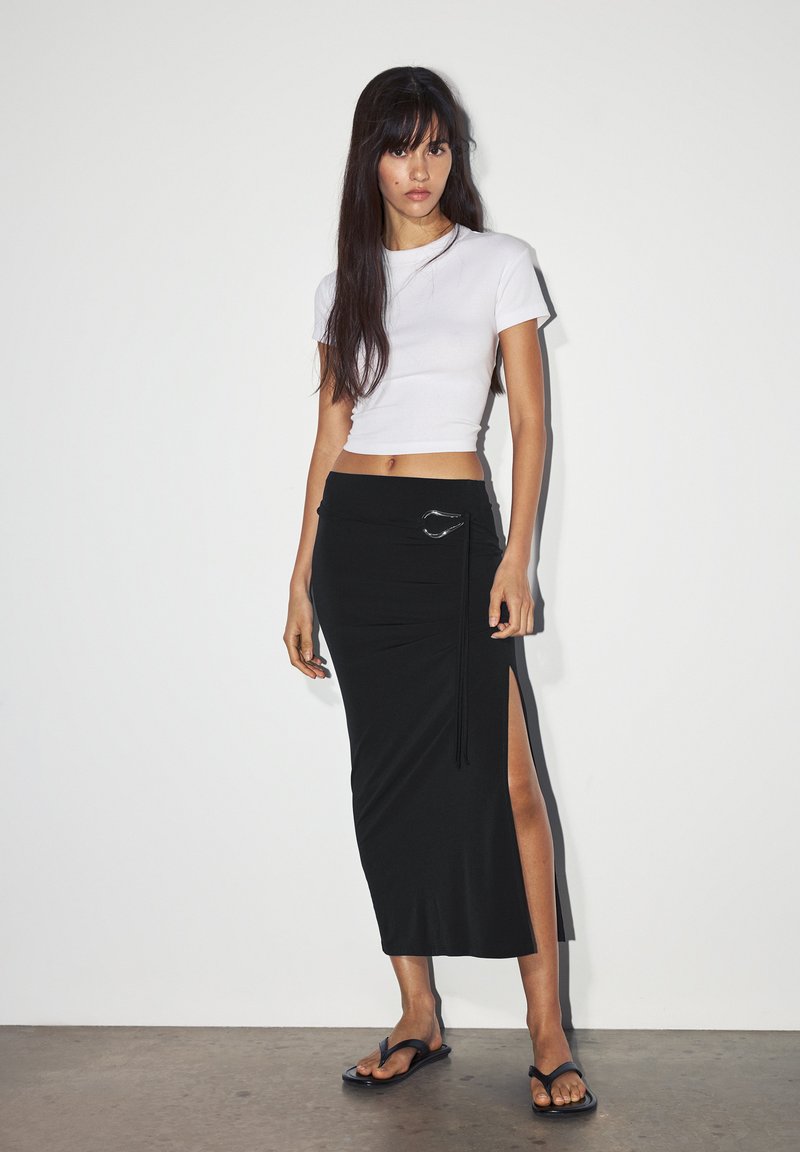 Black midi skirt with side slit and silver hardware accent, paired with a white short-sleeve crop top and black flip-flops on a neutral background.