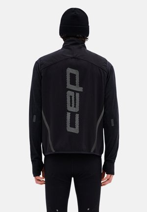 Persona che indossa un berretto nero, una maglia nera a maniche lunghe e un gilet nero con grande scritta riflettente "cap" sulla schiena, in piedi di spalle.