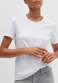 Vrouw in een eenvoudige witte korte mouwen T-shirt en grijze spijkerbroek, handen lichtjes samengevouwen ter hoogte van de taille, neutrale achtergrond.