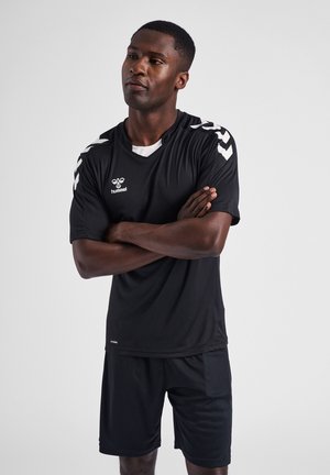 Man in zwarte sportkleding met witte chevronpatronen op de schouders, staande met gekruiste armen tegen een effen grijze achtergrond.