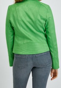 Donna che indossa una giacca verde brillante e jeans di denim grigi, vista da dietro, in piedi contro uno sfondo chiaro e semplice.