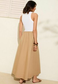 Top blanco sin mangas con textura acanalada, combinado con una falda maxi beige fluida. La modelo lleva accesorios y sandalias marrones.