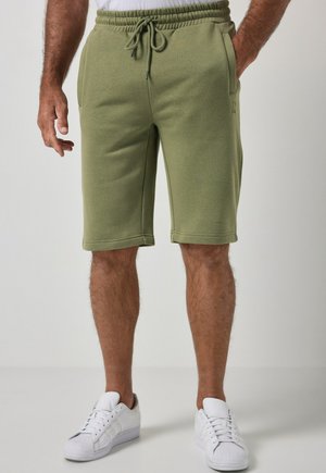 Homme portant un short vert olive à cordon et des baskets blanches, debout avec une main dans la poche devant un fond uni.