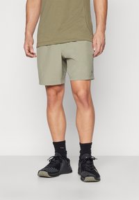 Pantalones cortos atléticos color caqui con una textura suave, que presentan un logo negro. Llevados con zapatillas negras de caña baja y calcetines tipo crew.
