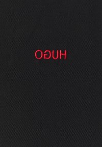 Černá texturovaná látka se červeně vyšitým logem "HUGO" našitým v obráceném směru uprostřed.
