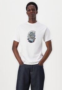 White cotton t-shirt featuring a graphic print in blue and black with the text "Santa Cruz." Paired with dark denim pants.