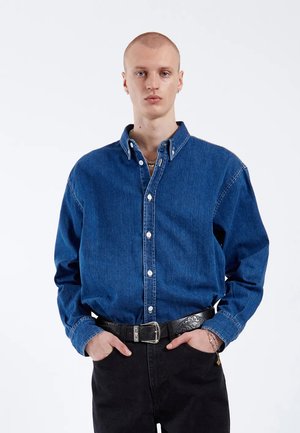 Dunkelblaues Jeanshemd mit Button-Down-Kragen, langen Ärmeln und weißen Knöpfen. Kombiniert mit schwarzen Hosen und einem strukturierten schwarzen Gürtel.
