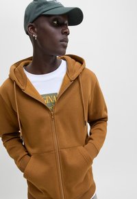 Ember barna cipzáras kapucnis pulóvert visel, kezét a zsebében tartja, fehér grafikás pólót és sötétzöld sapkát, oldalra néz egy egyszerű háttér előtt.