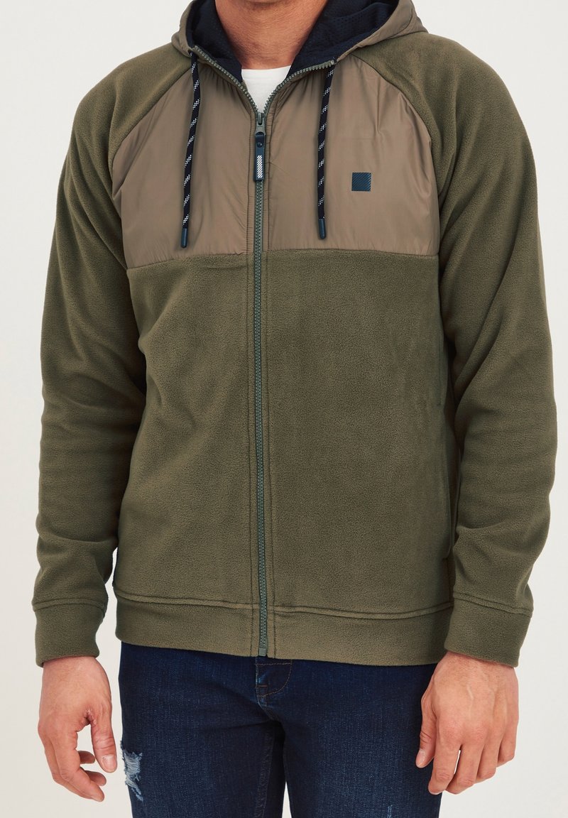 Homme portant un sweat à capuche zippé en polaire vert olive avec cordons sombres et panneau matelassé sur la poitrine, associé à un jean bleu foncé.