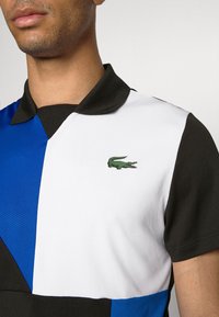 Pikétröja i svart, blått och vitt. Har en klassisk krage och en liten grön krokodil-logotyp på bröstet. Strukturerad tyg.