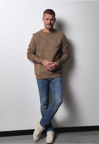 Brun sweatshirt med prægede logo, blå jeans og beige sneakers. Modellen står op ad en ensfarvet væg, med armene korslagt foran sig.
