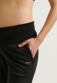 Zwarte sweatpants met een brede elastische tailleband, voorzien van een zijzak en een klein 'Woma'-logo in zilverdraad nabij de zak.