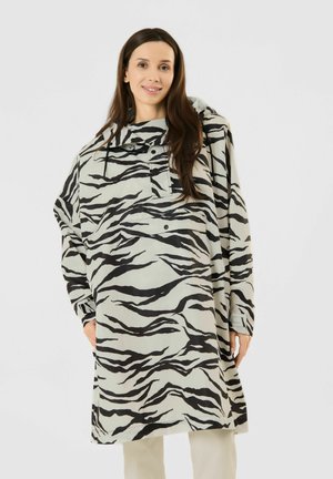 Woman wearing a loose, long-sleeve zebra-striped jacket with a hood, standing with hands in pockets, smiling at the camera on a white background.