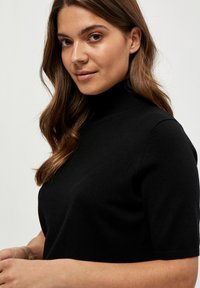Young woman with long brown hair wearing a black short-sleeve turtleneck sweater, looking slightly to the side, light background.