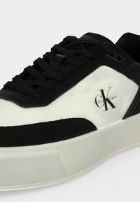 Черно-бял маратонка Calvin Klein с черни връзки и лого отстрани, с дебела бяла подметка на обикновен фон.