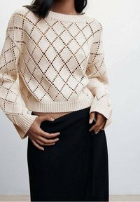 Pull en tricot écru avec motif en losange, doté de larges poignets et d'un ourlet court. Associé à une jupe noire.