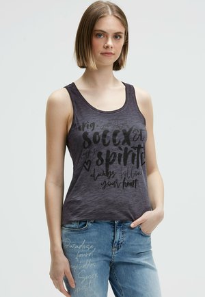 Dunkelgraues Tanktop aus weichem Material, mit kontrastierendem schwarzen Grafikslogan und einer lockeren Passform. Dazu trägt die Person blaue Jeans mit Schriftzügen.
