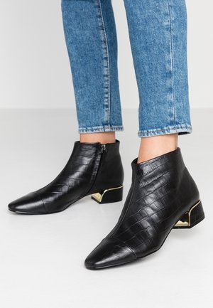 Botines negros con textura de cocodrilo, cierres laterales y tacones bajos metálicos, usados con jeans azules sobre un suelo claro y fondo liso.
