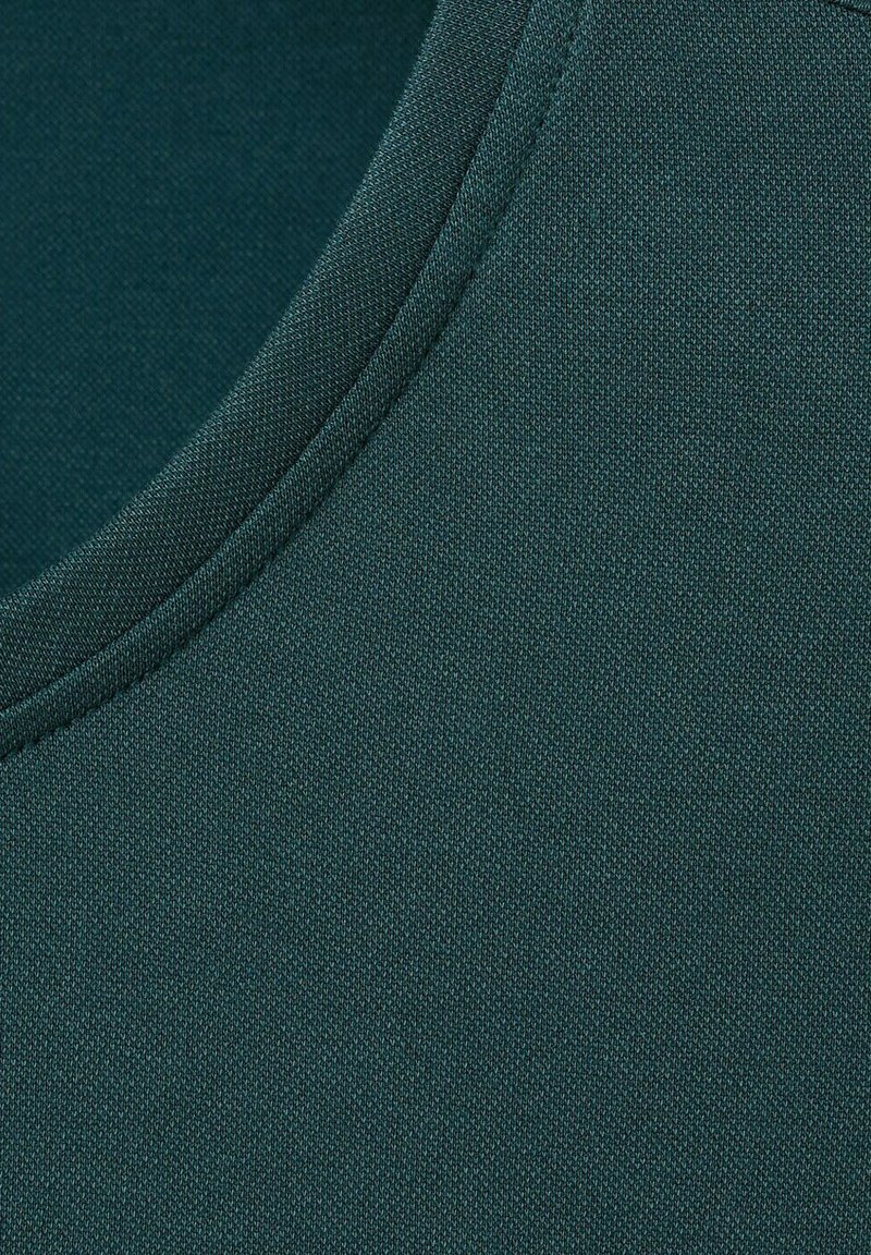 Tissu vert foncé avec une fine texture tricotée et un détail de couture courbe près du coin supérieur gauche.