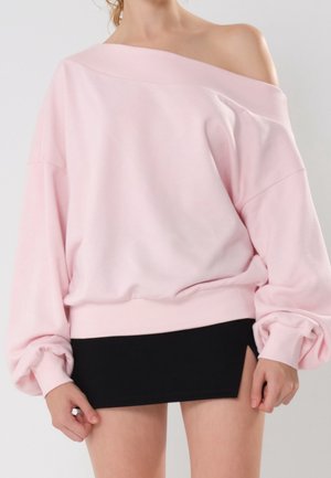 Femme portant un sweat-shirt ample rose clair à épaules dénudées avec des manches bouffantes et une jupe courte noire.