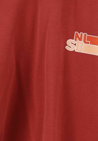 T-shirt en coton rouge avec un texte graphique blanc et orange « NL » et « SB » sur le devant. Texture douce avec des manches courtes.