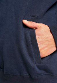 Navy blue sweatshirt featuring a side pocket; close-up shows soft texture and a hand resting in the pocket, emphasizing practicality and design.