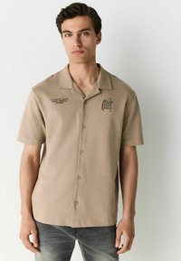 Beige short-sleeve shirt with a camp collar, featuring a button-down front, black printed graphics, and a smooth texture.