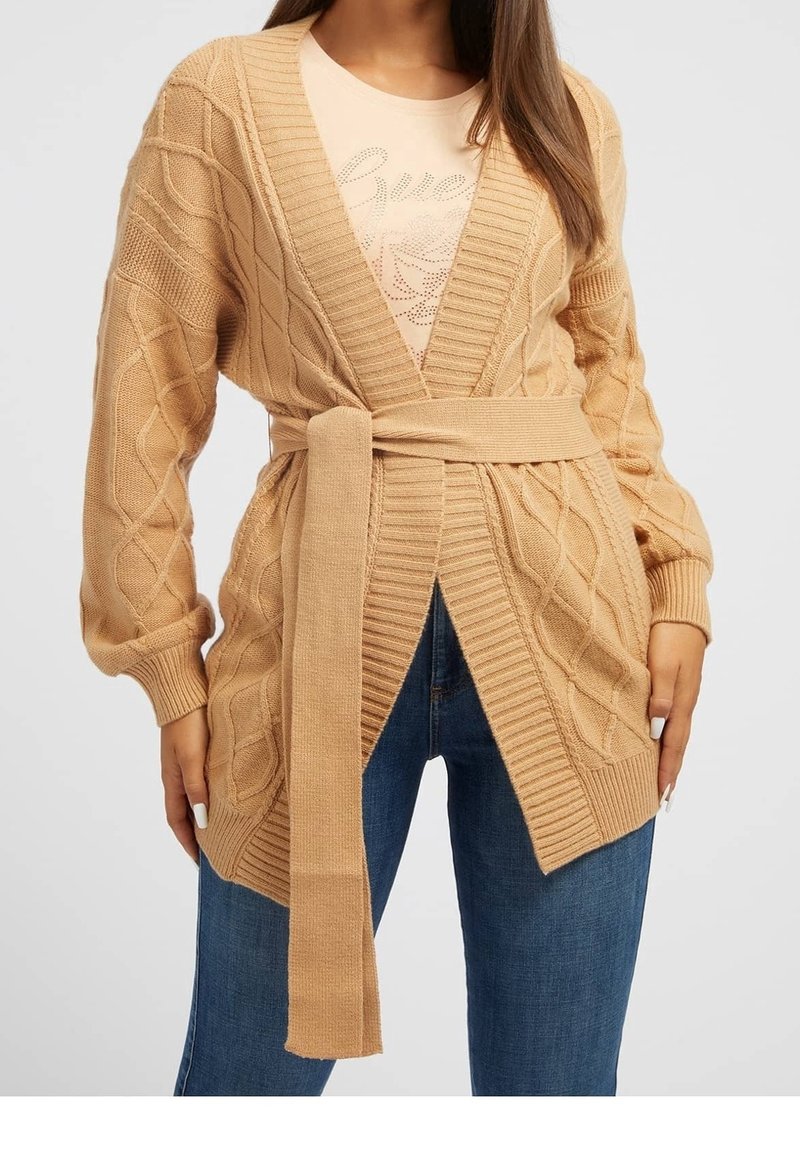 Cardigan beige en tricot avec un motif torsadé, col en V, manches longues et une ceinture à la taille. Porté avec un haut clair et un jean bleu.