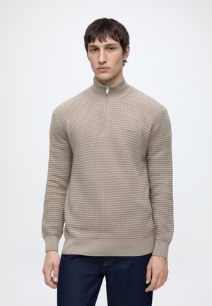 Homme portant un pull beige texturé à fermeture éclair partielle et un jean bleu foncé, debout devant un fond clair uni.
