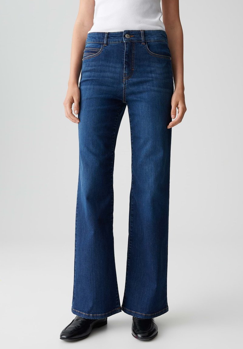 Donna che indossa jeans a vita alta svasati blu scuro con tasche frontali e mocassini neri, in piedi contro uno sfondo chiaro e uniforme.