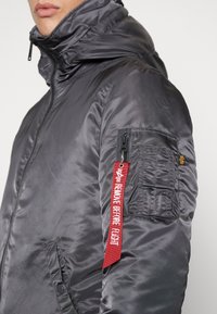 Gray insulated jacket with a high collar, visible stitches, and a zippered pocket. Features a red tag stating "REMOVE BEFORE FLIGHT."