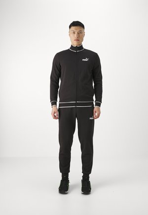 Tuta nera realizzata in tessuto morbido; giacca con zip e accenti bianchi, pantaloni abbinati e scarpe nere con dettagli verdi. Logo Puma visibile.